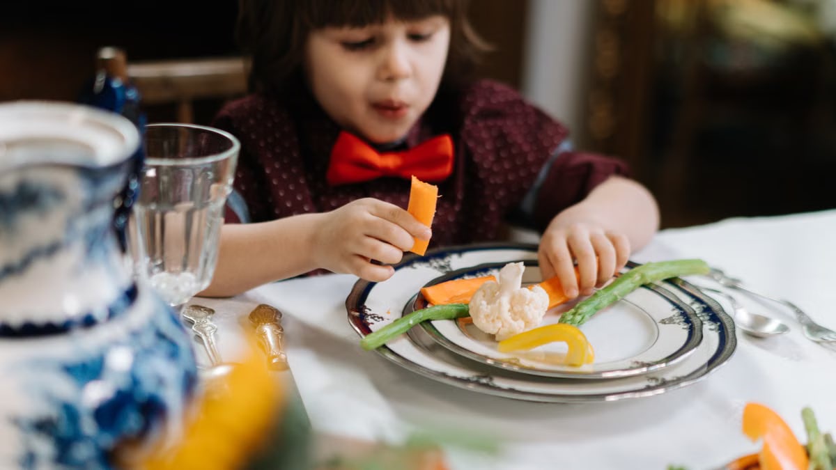 A child in a red bow tie enjoys a healthy meal with vegetables indoors.